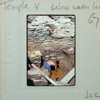 A man working on a stone structure below water level, with the photo labeled as Temple V.
