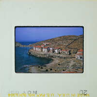 A vintage photo of a coastal village with houses and a pier on the water's edge, under a blue sky.