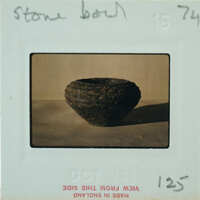 The image shows an old, weathered stone bowl with a rough texture, placed on a surface and photographed against a neutral background.