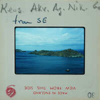 The image shows a vintage film strip with a photograph of an island and sea, accompanied by handwritten notes indicating the name "Nikos" and the location as "SE". The film strip is part of a collection or exhibit, as suggested