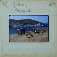 A vintage photo of people standing on a rocky shore near the water, with mountains and houses in the background.