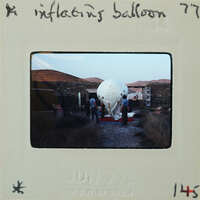 A vintage photograph of a group of people standing around an inflated balloon on a dirt ground with rocky hills in the background.