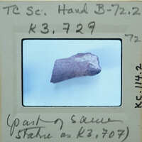 A small, dark rock specimen on a white surface with labels and handwritten notes around it.