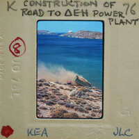 A photograph of a dirt road construction site with a body of water and mountains in the background, presented on a vintage film strip.