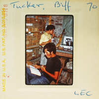 Two men sitting in a room with boxes, one looking at a book and the other drinking from a cup.