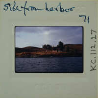 A vintage photograph of a serene landscape with water, hills, and trees, featuring an old building on the shore.