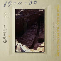 A photograph of an ancient cave entrance with stone walls and a small stream, taken from a low angle.