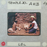 A woman kneeling on the ground near a stone structure, possibly an archaeological site.