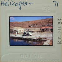 A helicopter parked on a dock next to a body of water, with mountains in the background.
