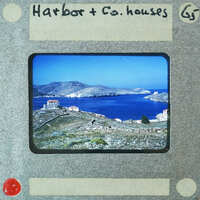 A vintage photo of a harbor with houses overlooking the sea and mountains in the background.