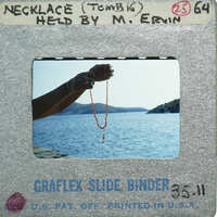 A person holding a necklace over water, with mountains in the background.