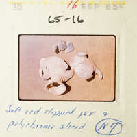 The image shows a collection of broken pottery shards on a table, with a focus on a red-rimmed shard and a small piece of red clay.