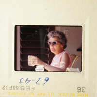 A vintage photo of an older woman holding a cup, sitting on a porch with her sunglasses on.