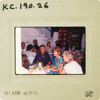 A vintage photo of a group of people sitting around a table, possibly at a social gathering or event. Catalog number KC.190.26