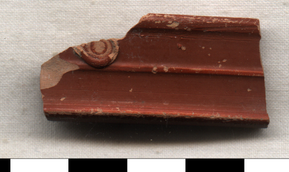 Fragment of terra sigillata (Roman red-slipped pottery) showing a profile molding with decorative relief element, likely part of a vessel rim or base with characteristic reddish-brown slip and raised decorative details.